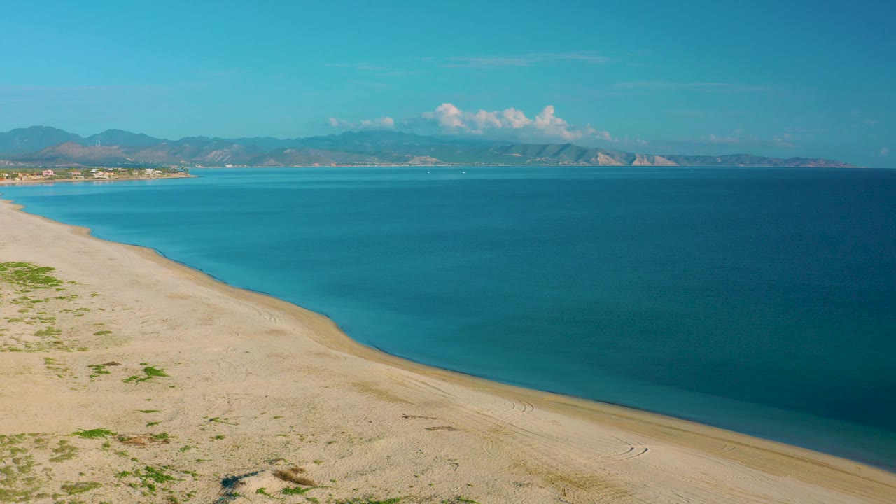 A high coastal view in La Ribera shows pale beach along the left and bottom edges, blue bay water across the frame, mountains in the distance, and no visible people.