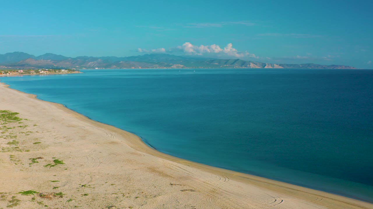 La Ribera beach fills the lower frame with sparse vegetation and sand patterns, while broad blue water and a distant mountain coastline sit beyond, with no visible people.