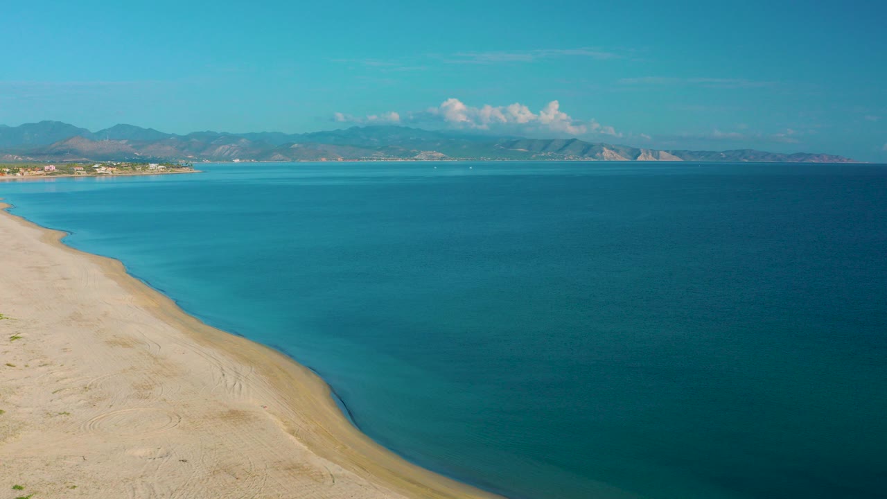 Opening aerial frame over La Ribera, Baja California Sur, with pale sand at left, blue sea across the frame, distant mountains under scattered clouds, and no visible people.