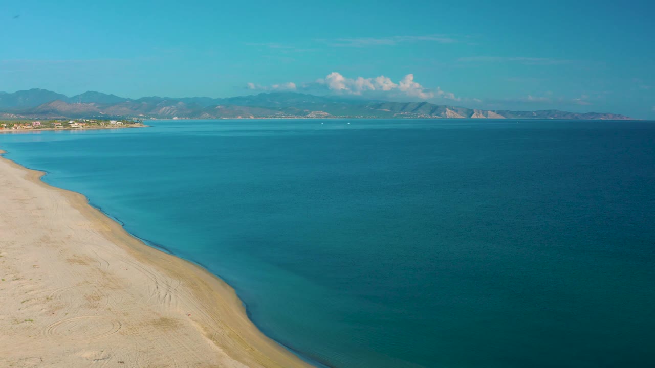 Wide aerial view in La Ribera with the beach tapering along the left side, deep blue open sea to the right, hazy mountains on the horizon, and no visible people.