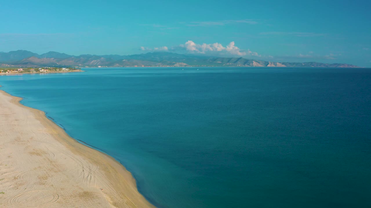 Aerial frame over La Ribera where turquoise shallows meet darker blue water beside the sandy beach, with mountain ridges and white clouds in the background and no visible people.