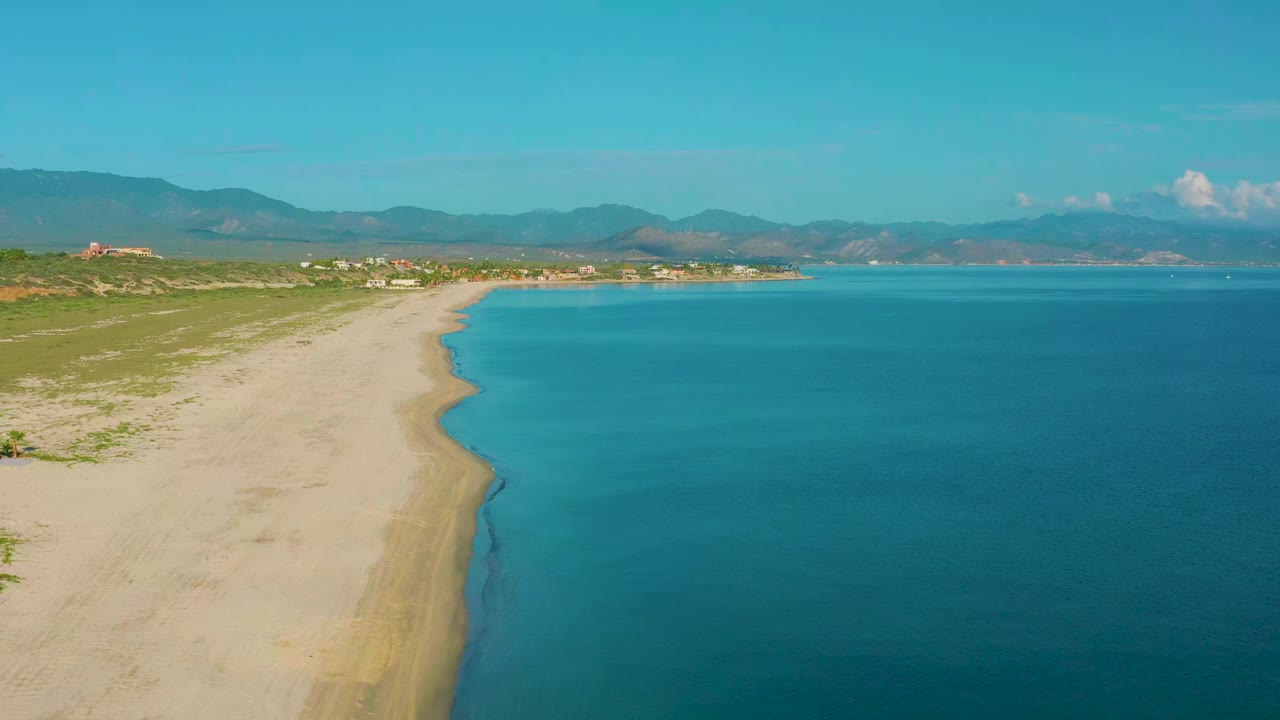Opening aerial frame over La Ribera, Baja California Sur shows a sandy beach curving beside turquoise sea, low coastal brush, distant buildings, mountains, and no visible people.