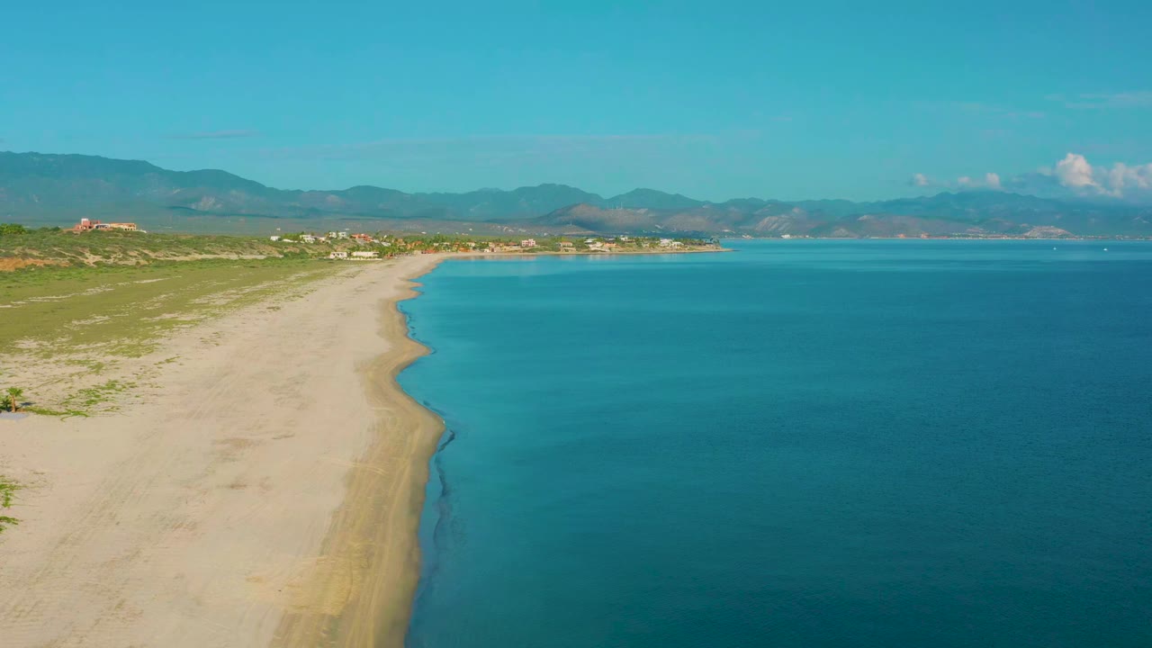 At 0:01 the La Ribera shoreline remains wide in frame, with the beach edge slightly farther along the left side, blue water to the right, distant buildings, mountains, and no visible people.
