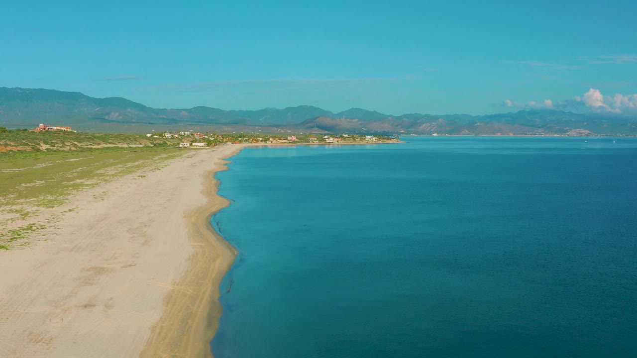 Opening frame over La Ribera, Baja California Sur shows pale sand curving beside turquoise water, low green scrub, distant buildings, mountains, and no visible people.