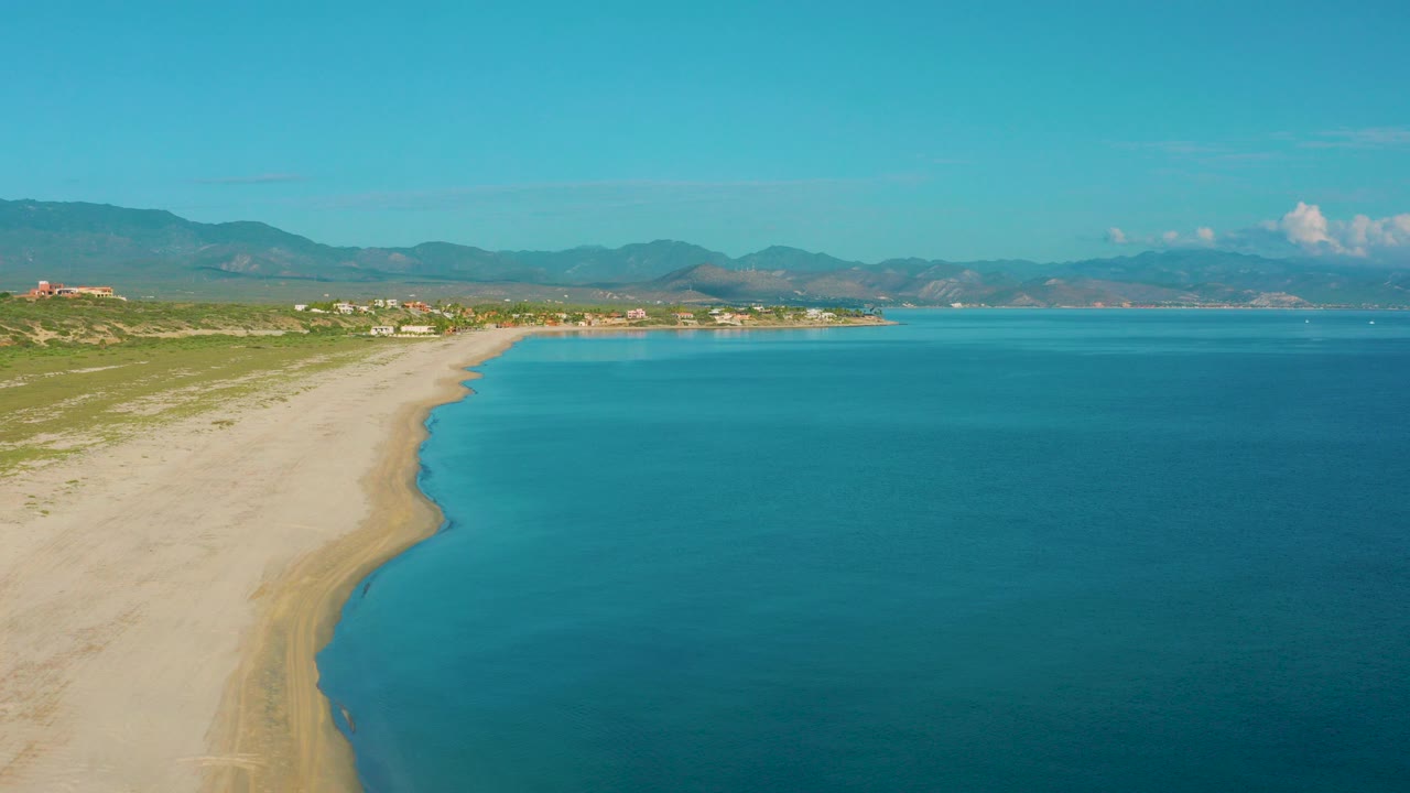 Aerial view of La Ribera, Baja California Sur, with a pale sandy beach curving beside clear blue water, low surf near shore, distant mountains, and no visible people.