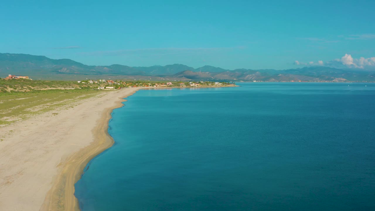 Aerial view of La Ribera coastline with the beach running diagonally through frame, calm blue sea filling the right side, distant settlement, mountains, and no visible people.