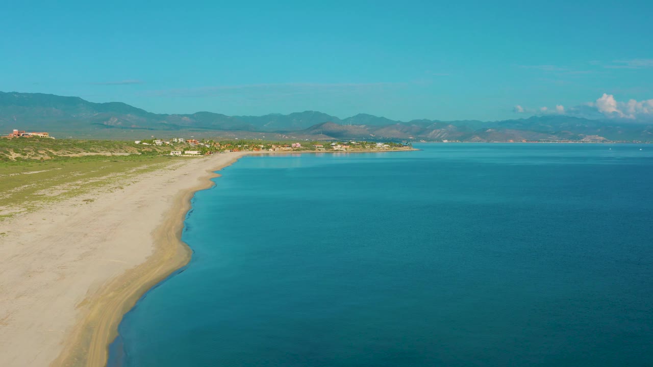 Aerial shoreline with pale sand, clear water, coastal scrub, and mountains.