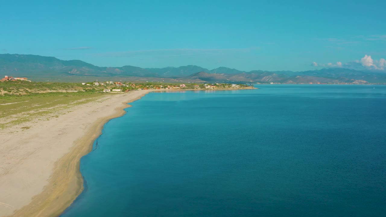 Aerial frame over La Ribera with the sandy shoreline forming a diagonal from lower left to midframe, clear blue water filling the right side, distant settlement, mountain backdrop, and no visible people.