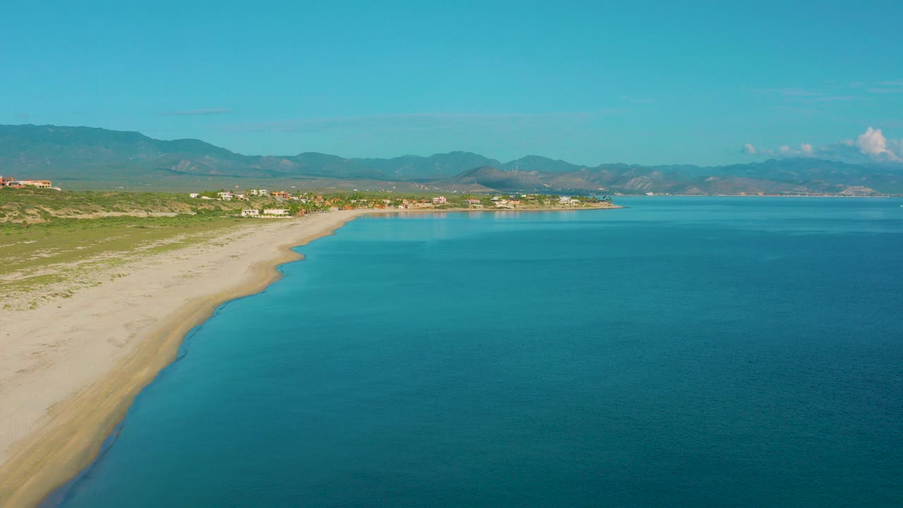 Wide aerial frame over La Ribera, Baja California Sur, with blue ocean on the right, pale sandy shoreline on the left, distant buildings and mountains, and no visible people.