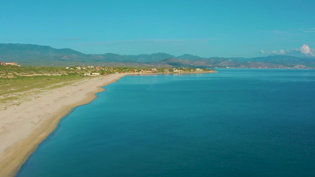 Aerial view of La Ribera with open blue water occupying most of the frame, a pale beach along the left, scrub and distant structures inland, hazy mountains, and no visible people.
