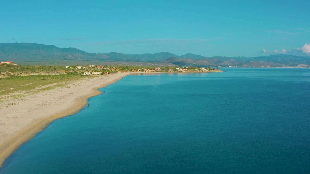 Aerial view over La Ribera, Baja California Sur, with blue sea in the foreground, pale sand curving along the left side, green coastal vegetation, distant mountains, and no visible people.