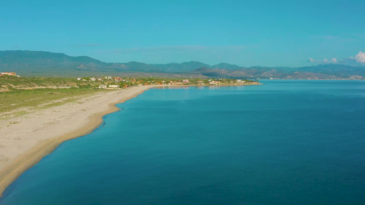 Wide aerial frame from La Ribera, Mexico, showing turquoise water across the foreground, a pale sandy shore on the left, green dunes, small coastal buildings, mountain ridges, and no visible people.