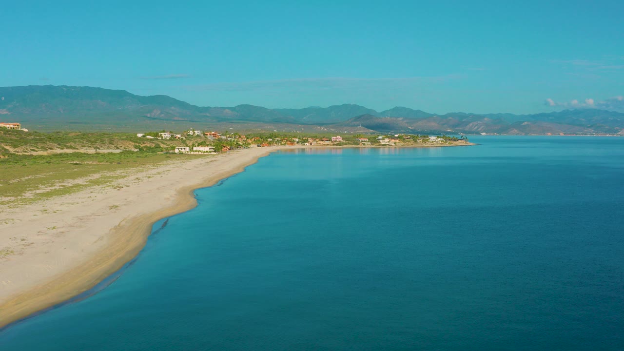 Aerial view over La Ribera, Baja California Sur, with pale sand curving along bright blue water, low vegetation, distant buildings, mountains, and no visible people.