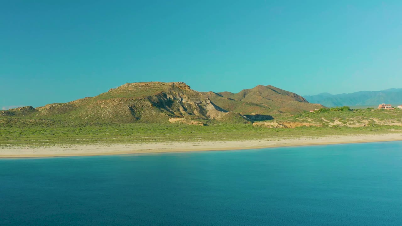 La Ribera shoreline frame with calm blue water below, a pale beach band across the center, scrubland and brown rocky hills beyond, and no visible people.