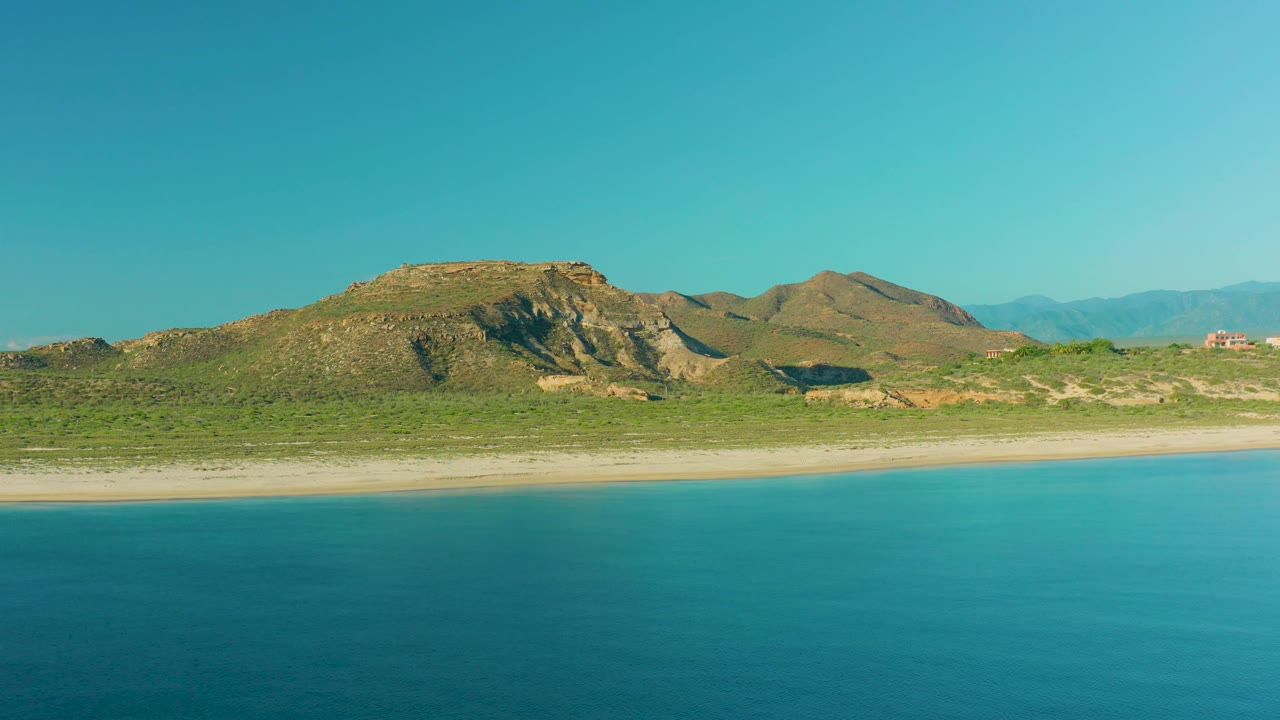 A wide La Ribera coastal view shows turquoise water with subtle surface texture, an empty sand strip, green scrub, layered rocky hills, and no visible people.