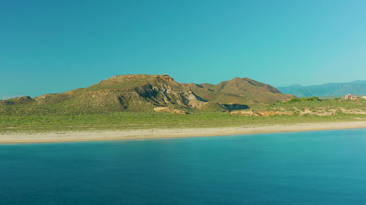 Blue water foreground, pale beach, scrubland, and rocky coastal hills.