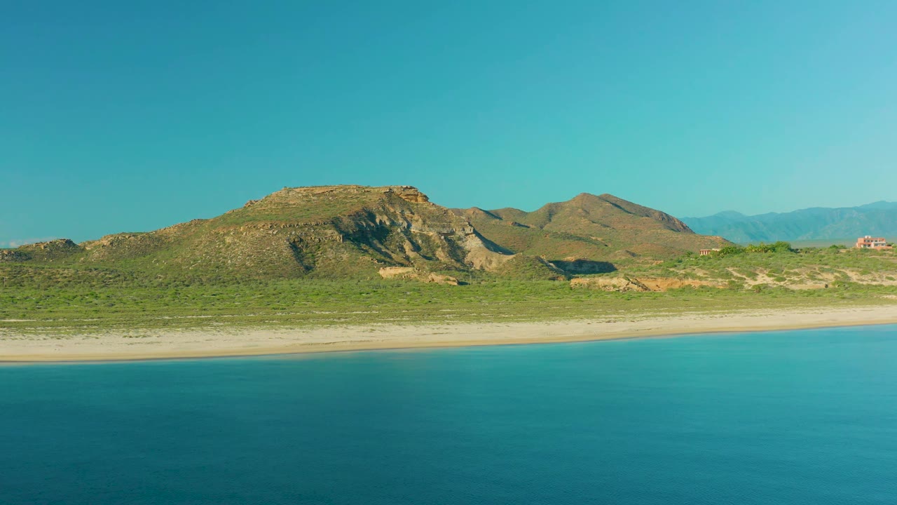 At 0:01, the La Ribera coastline remains wide in frame, with turquoise water below, pale sand near the shore, green scrubland, rocky hills, and no visible people.