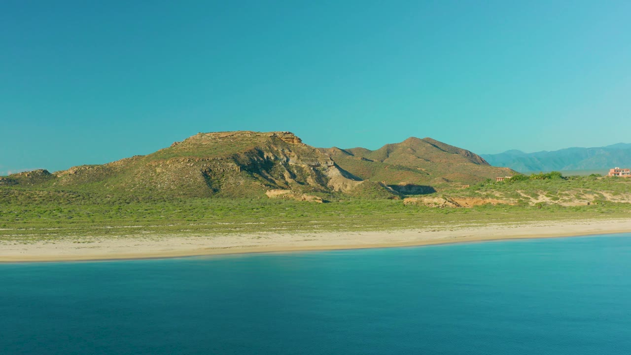 At 0:03, the aerial view shows calm blue water, a narrow sand strip, low green vegetation, layered tan hills, hazy distant ridges, and no visible people.