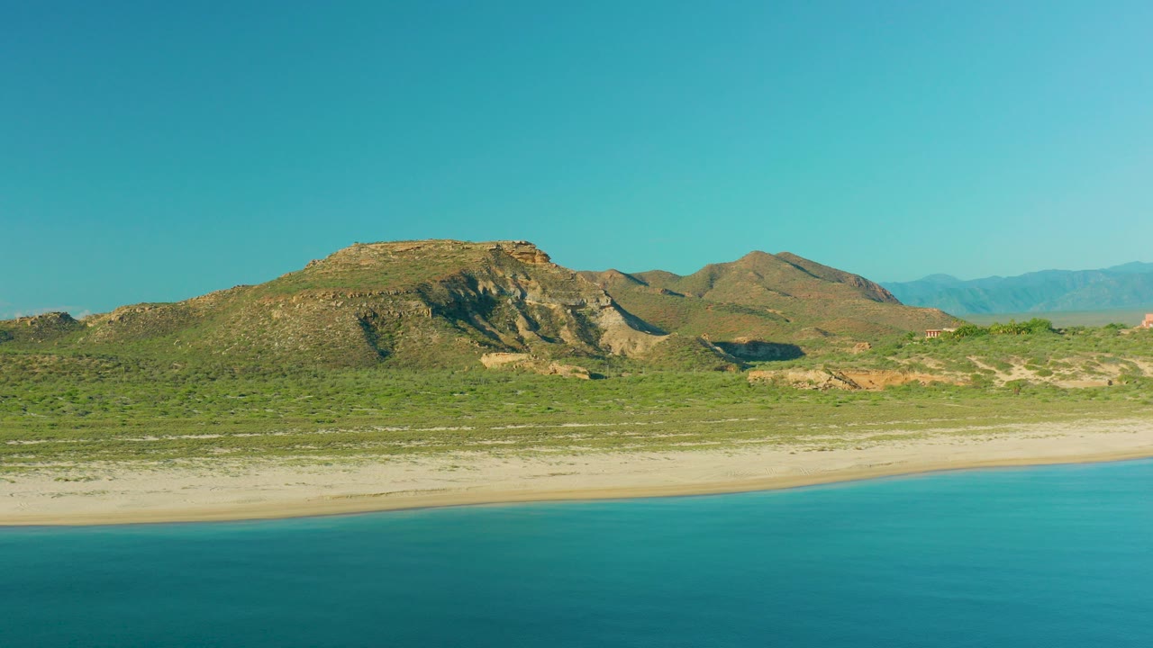At 0:00, a wide aerial frame shows turquoise water below a pale sand beach, scrub-covered land, rocky hills, distant mountains, and no visible people in La Ribera, Baja California Sur.