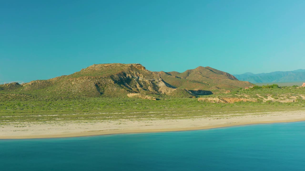 At 0:01, blue water runs along the lower frame while the beach, green scrub, tan rocky hills, faint background mountains, and no visible people define the La Ribera coastal view.