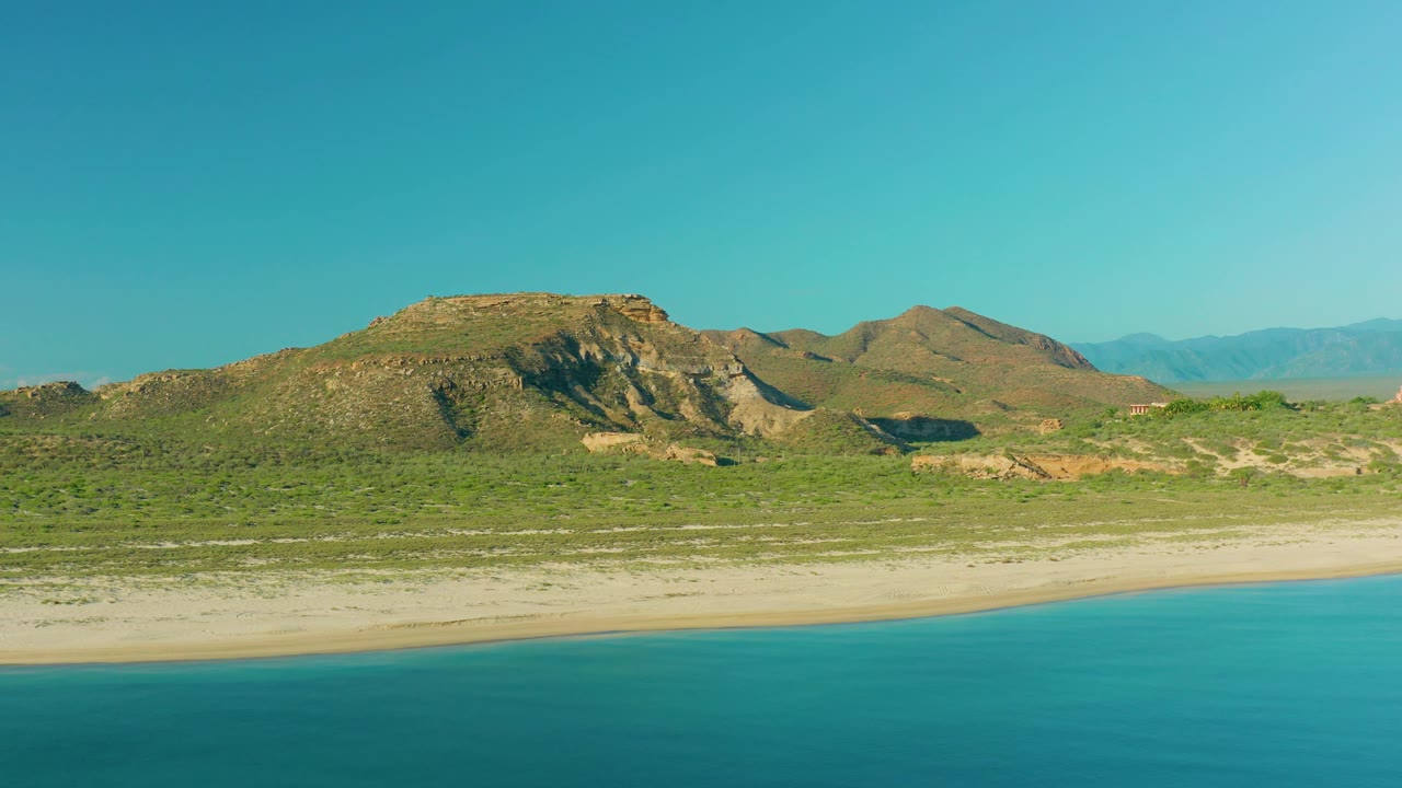 At 0:02, the aerial composition shows a turquoise band of sea at the bottom, a narrow pale beach, scrubland, brown hills, hazy mountains, and no visible people in La Ribera.