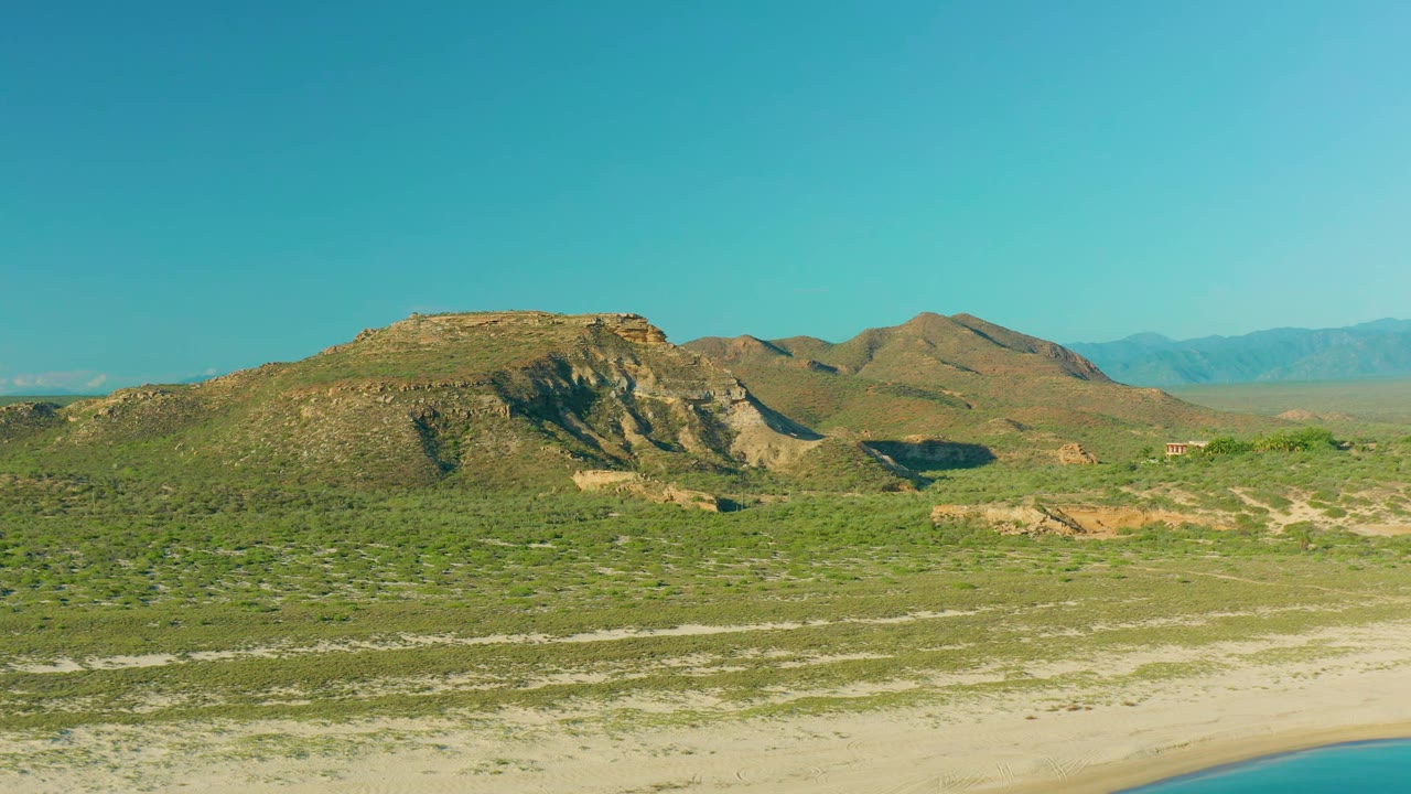 Wide aerial view in La Ribera, Baja California Sur with scrubland leading to low hills, distant blue mountains, a clear sky, a small water edge, and no visible people.