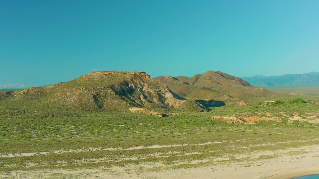 Aerial landscape in La Ribera, Baja California Sur with low rocky hills, green scrub, pale tracks or sandy patches, distant mountains, and no visible people.