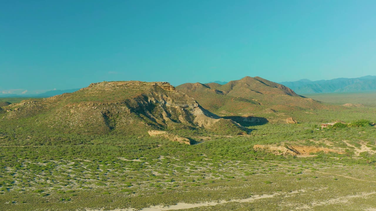 A broad scrub plain leads toward a rocky hill in La Ribera, Baja California Sur, Mexico, with pale dirt lines crossing the foreground and no visible people.