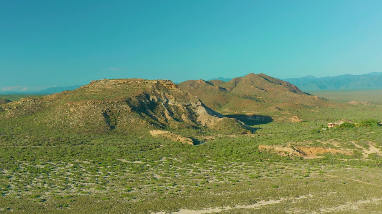 Green-brown vegetation fills the foreground below rocky hills in La Ribera, Baja California Sur, Mexico, with a small structure-like shape near the hillside and no visible people.