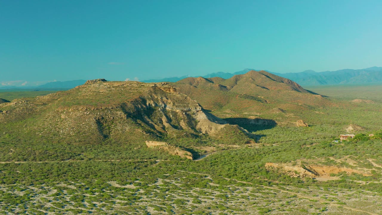 Opening aerial frame over La Ribera, Baja California Sur, showing green desert scrub, rocky hills, distant blue mountains, and no visible people.