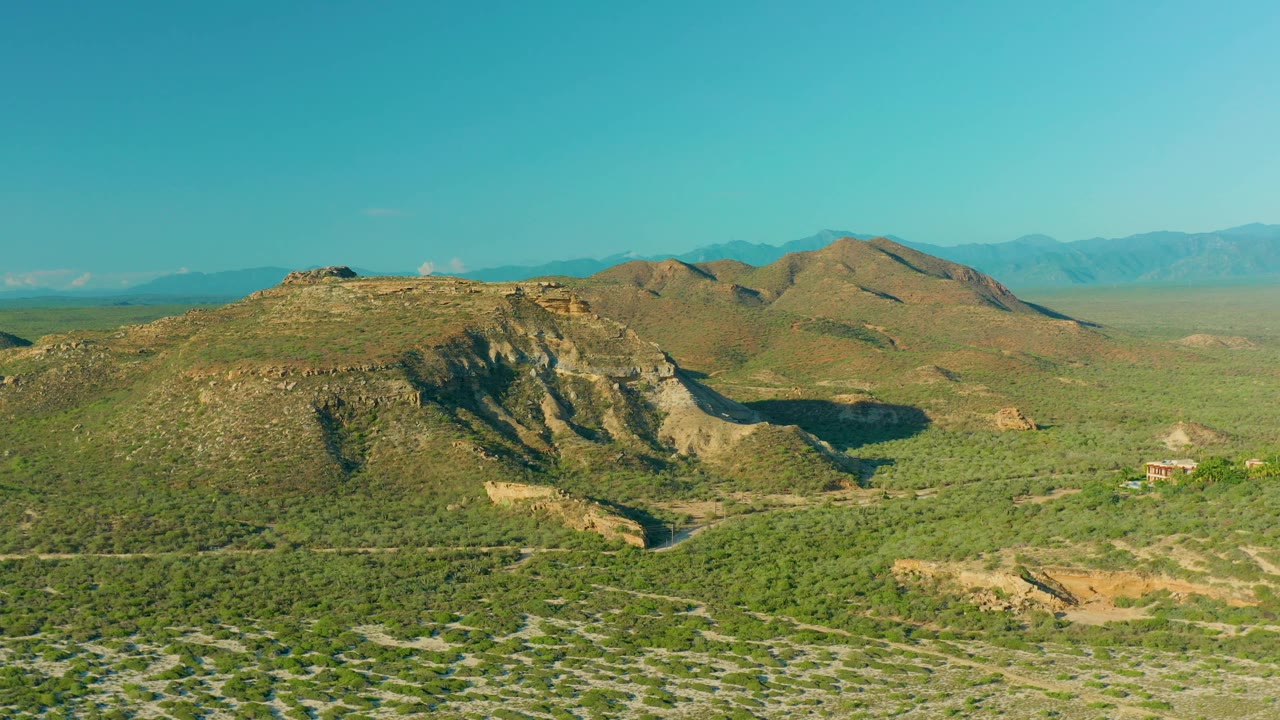 La Ribera aerial still with scrubland filling the lower frame, brown hills through the center, blue mountains in the distance, and no visible people.