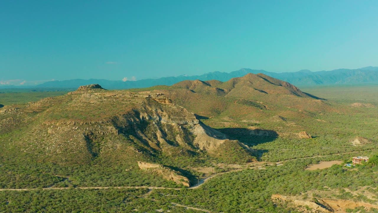 Aerial view over La Ribera, Baja California Sur, showing green scrubland, rocky hills, distant mountain ridges, and no visible people.