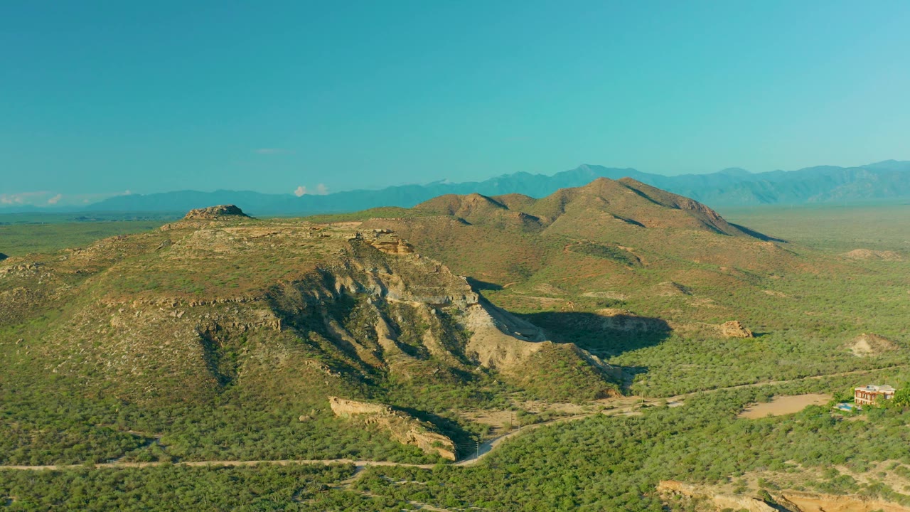 Wide aerial view of La Ribera, Baja California Sur, with scrub-covered hills, a pale road crossing the lower frame, hazy mountains in back, and no visible people.