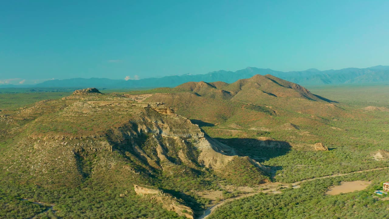 A slightly later aerial frame in La Ribera shows the rocky hill more centered, with green lowland brush, far blue mountains, and no visible people.
