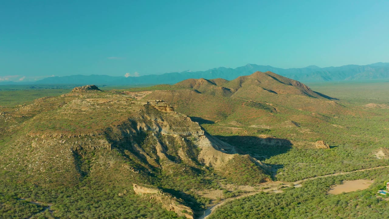 Aerial landscape in La Ribera with sunlit green hills, a pale dirt road crossing the lower frame, distant mountains, and no visible people.