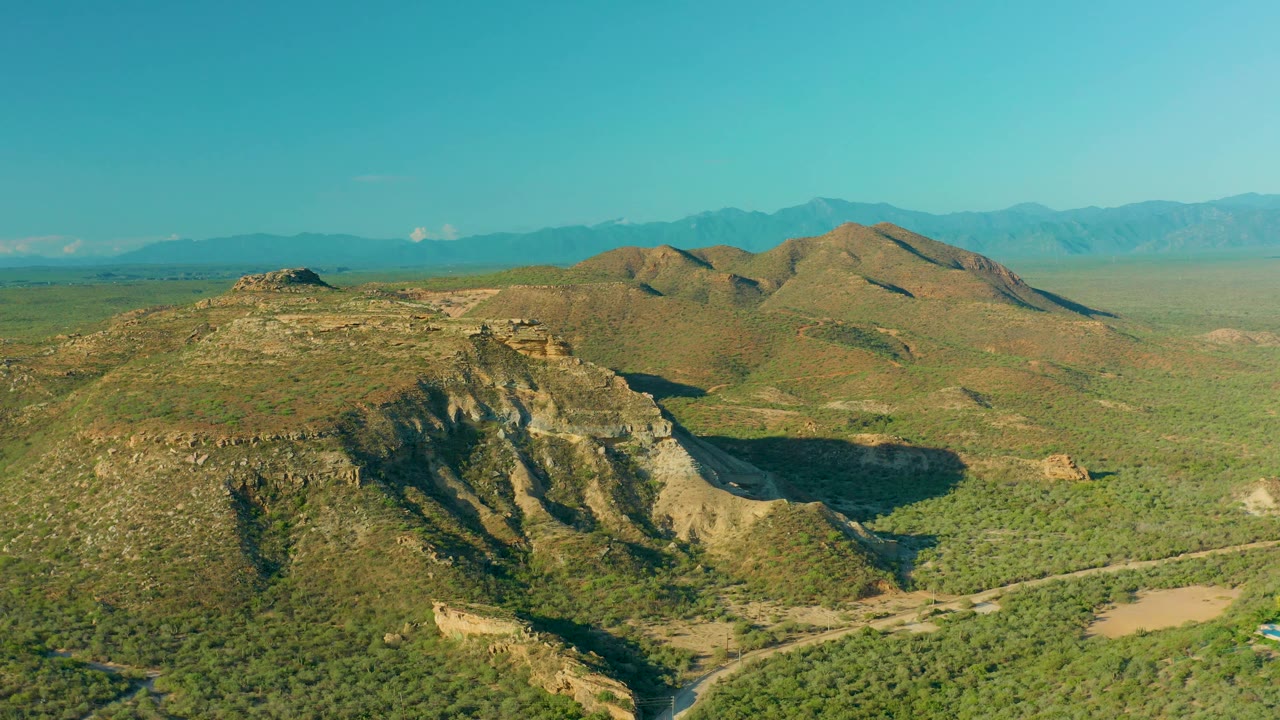 Wide aerial frame over La Ribera showing sunlit scrub hills, a deep shadow on the near ridge, a broad valley, and no visible people.