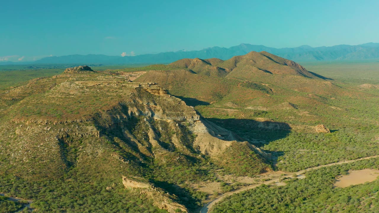 High aerial frame over La Ribera, Baja California Sur, showing the ridge slightly farther across frame, a dirt road below, blue mountain horizon, and no visible people.