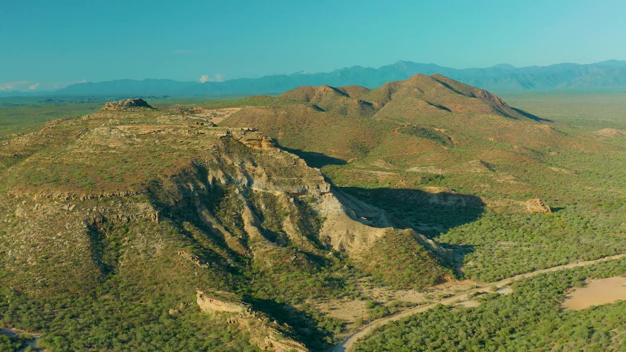 Wide aerial frame over La Ribera, Baja California Sur, with sunlit hills, darker slope shadows, scattered scrub, dirt tracks, distant ridges, and no visible people.