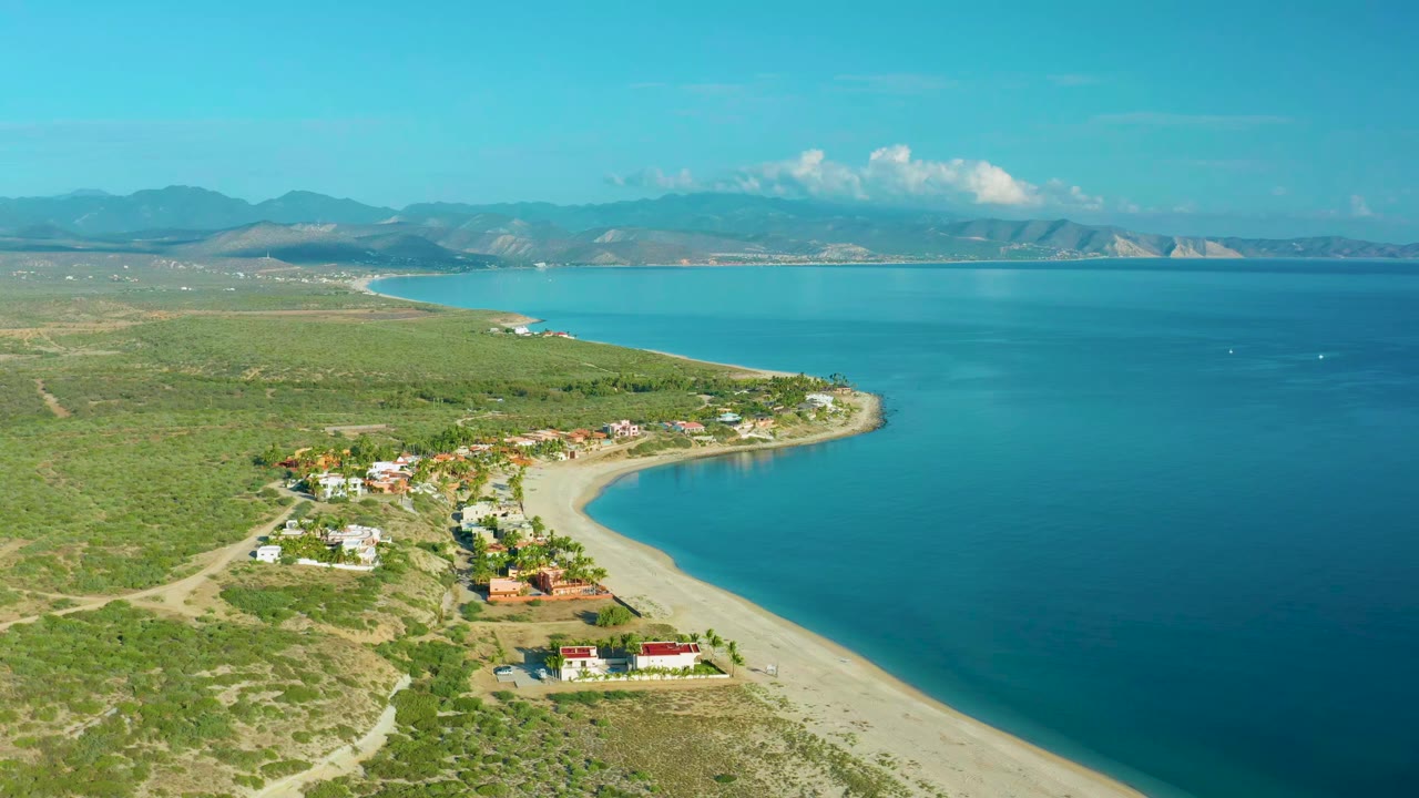 Wide aerial frame over La Ribera showing beach homes near the lower edge, a pale shoreline wrapping into calm blue water, distant mountains, and no visible people.