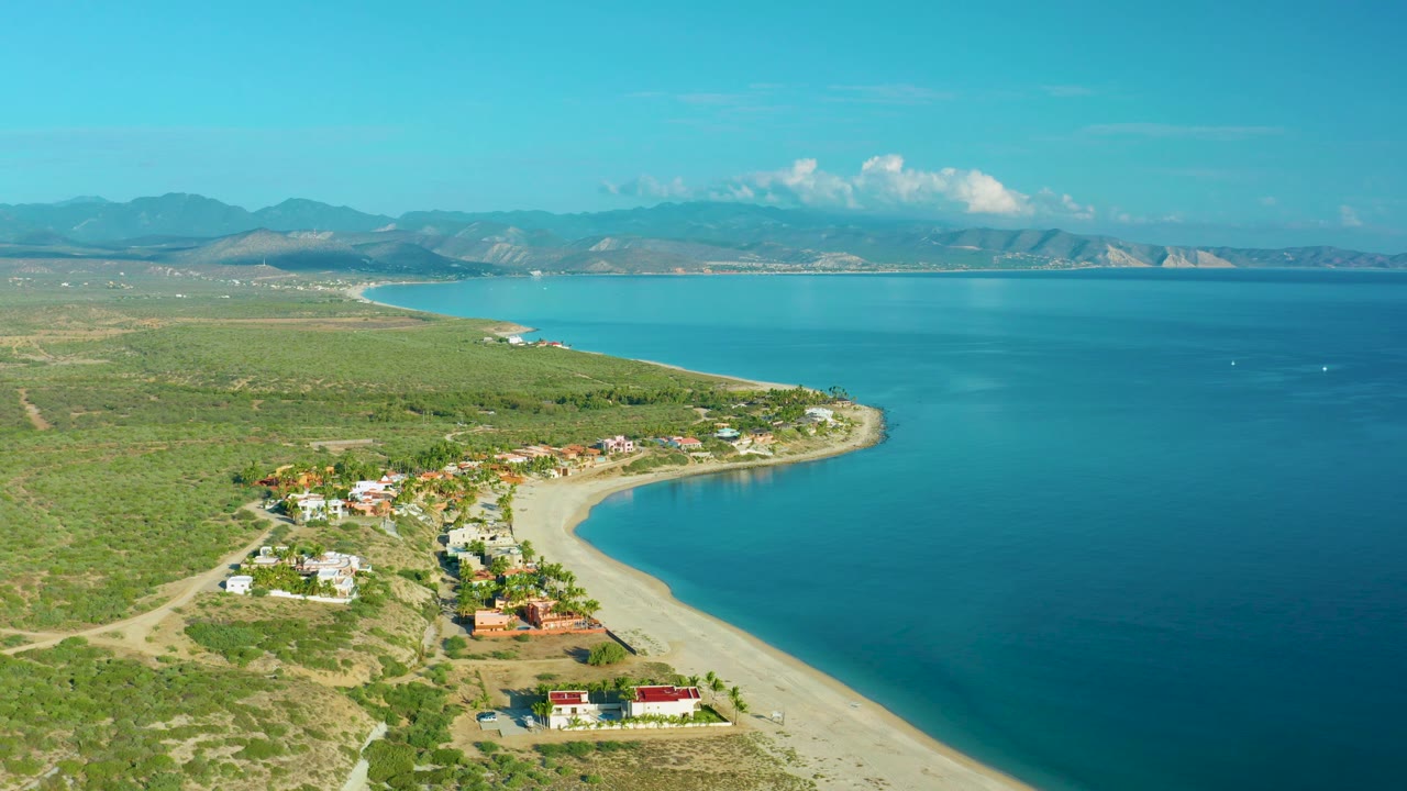 High aerial frame over La Ribera, Baja California Sur, with sandy beach sweeping around a point, houses near the water, dry green-brown vegetation inland, blue sea, distant mountains, and no visible people.