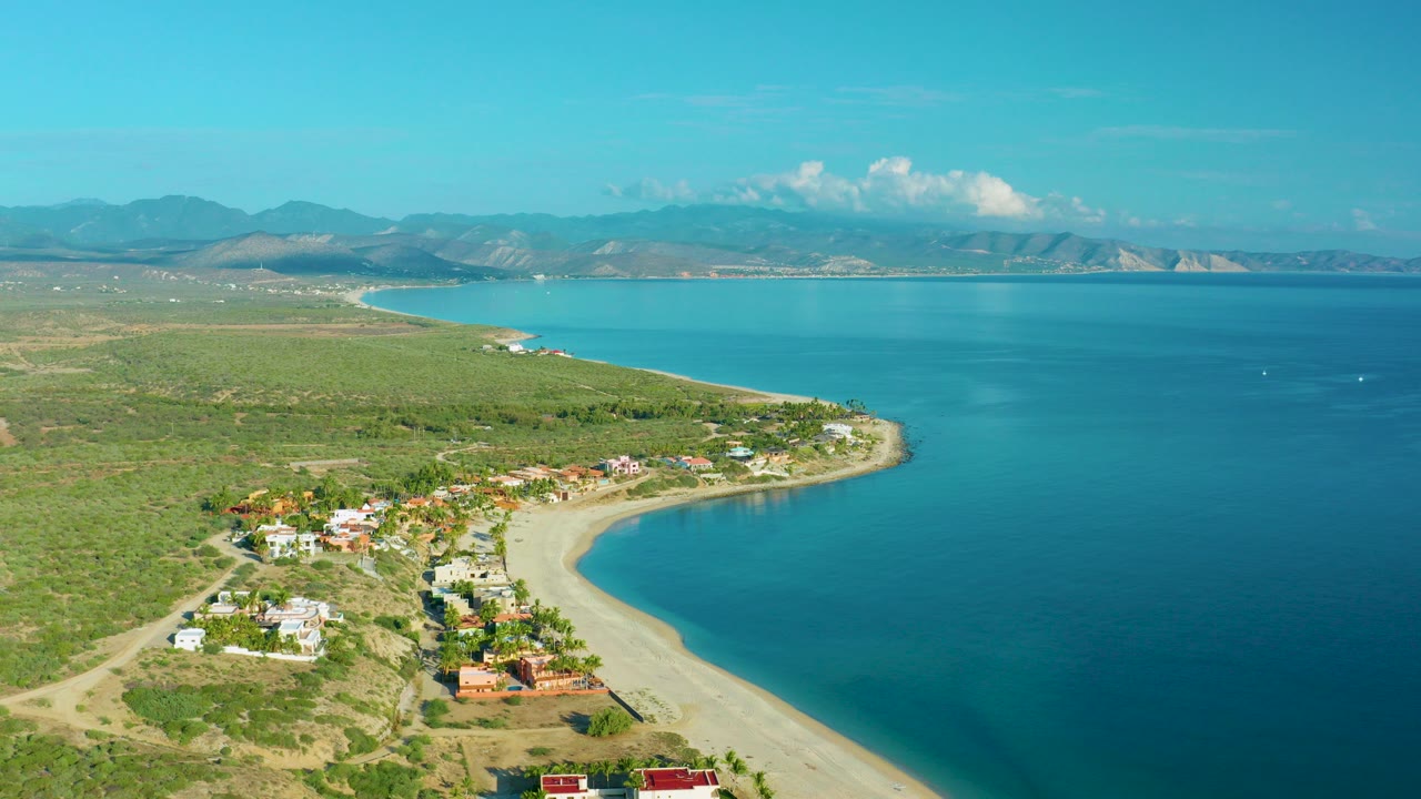 Aerial frame over La Ribera, Baja California Sur showing a curved pale beach, scattered homes and palms near shore, wide blue bay water, distant mountains, and no visible people.
