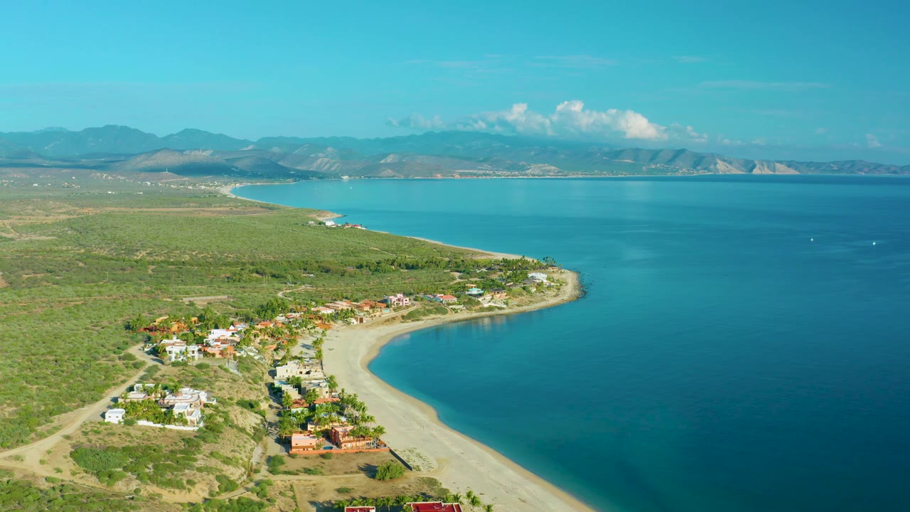 Aerial view in La Ribera, Baja California Sur looking over turquoise bay water beside a sandy curve, with scattered shore homes, open scrubland, distant mountains, and no visible people.