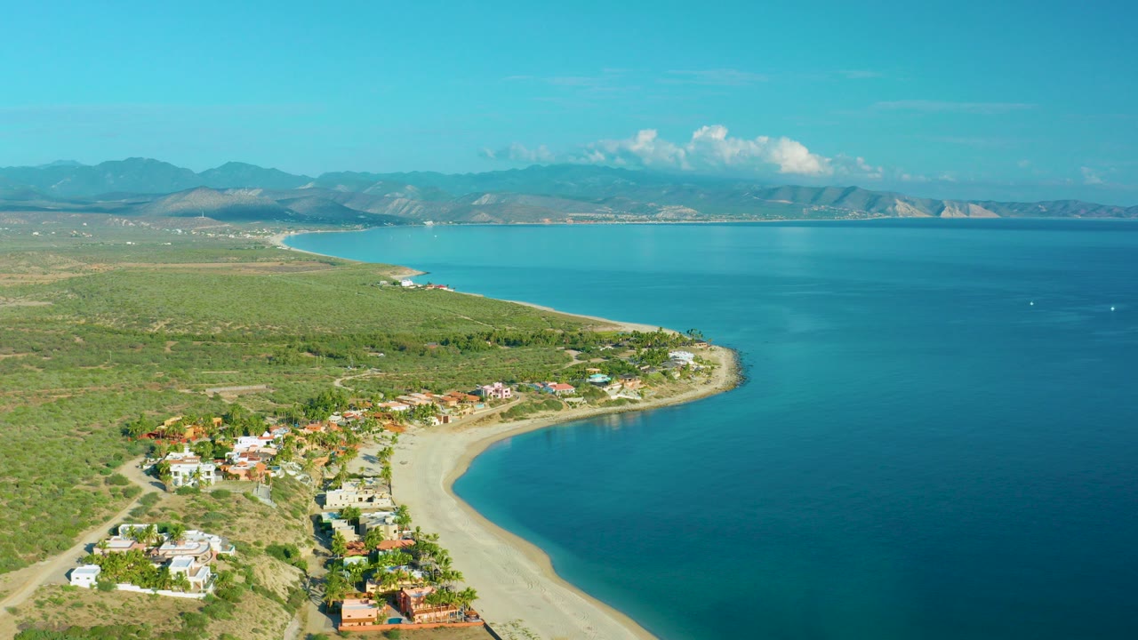 Aerial view over La Ribera, Baja California Sur showing coastal homes beside a curved sand beach, turquoise bay water, distant mountains, and no visible people.