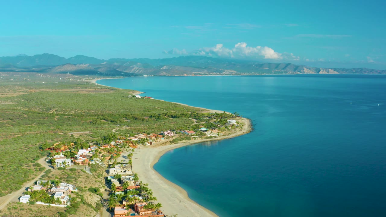 Aerial coastline view over La Ribera, Baja California Sur showing a beach cove, clustered houses, open bay water, hazy mountains, and no visible people.