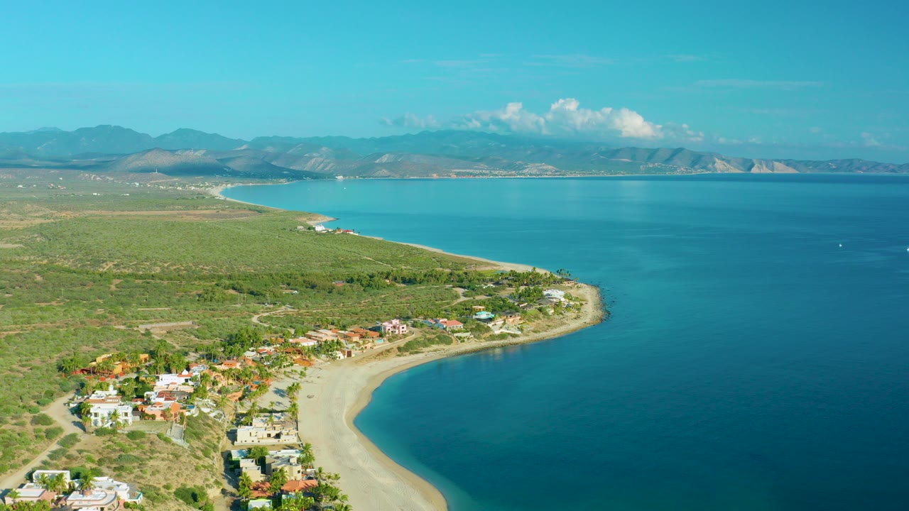 Opening aerial frame over La Ribera, Baja California Sur, with beach homes at the lower left, a curved sandy shore, blue bay water, distant mountains, and no visible people.