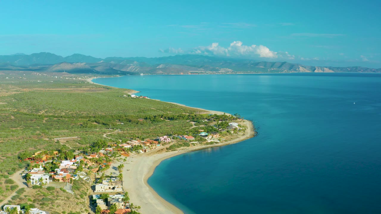 Wide aerial of beach homes, sandy shoreline, green scrubland, blue water, and mountains.