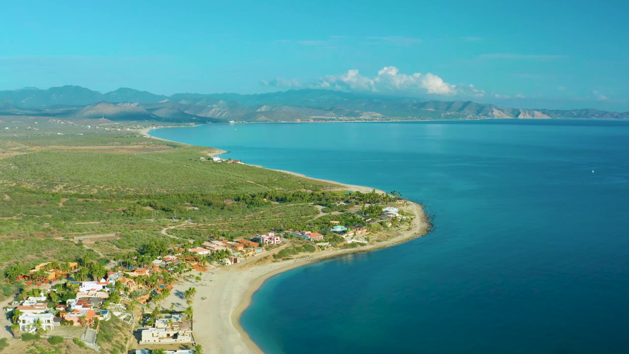 Aerial frame over La Ribera with shoreline homes at the lower left, a green coastal plain, deep blue bay water, distant mountains, and no visible people.
