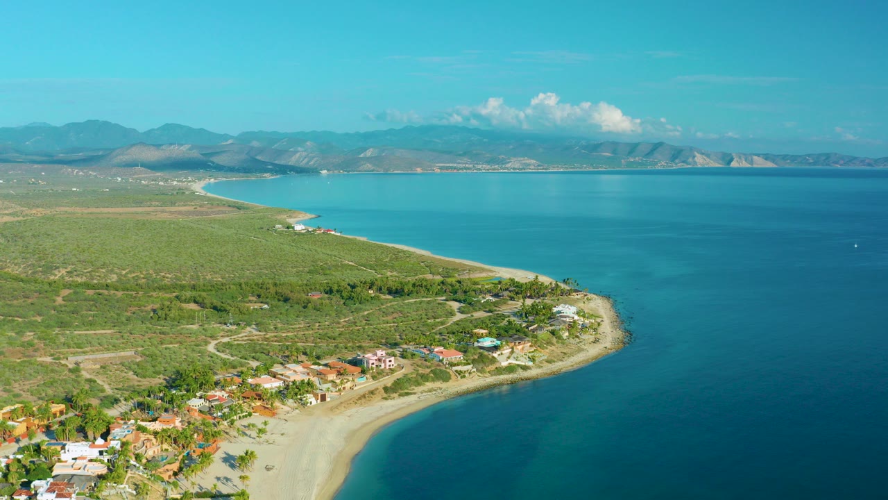 Aerial frame over La Ribera, Baja California Sur showing a curved sandy beach, blue sea, green scrubland, low coastal homes, distant mountains, and no visible people.