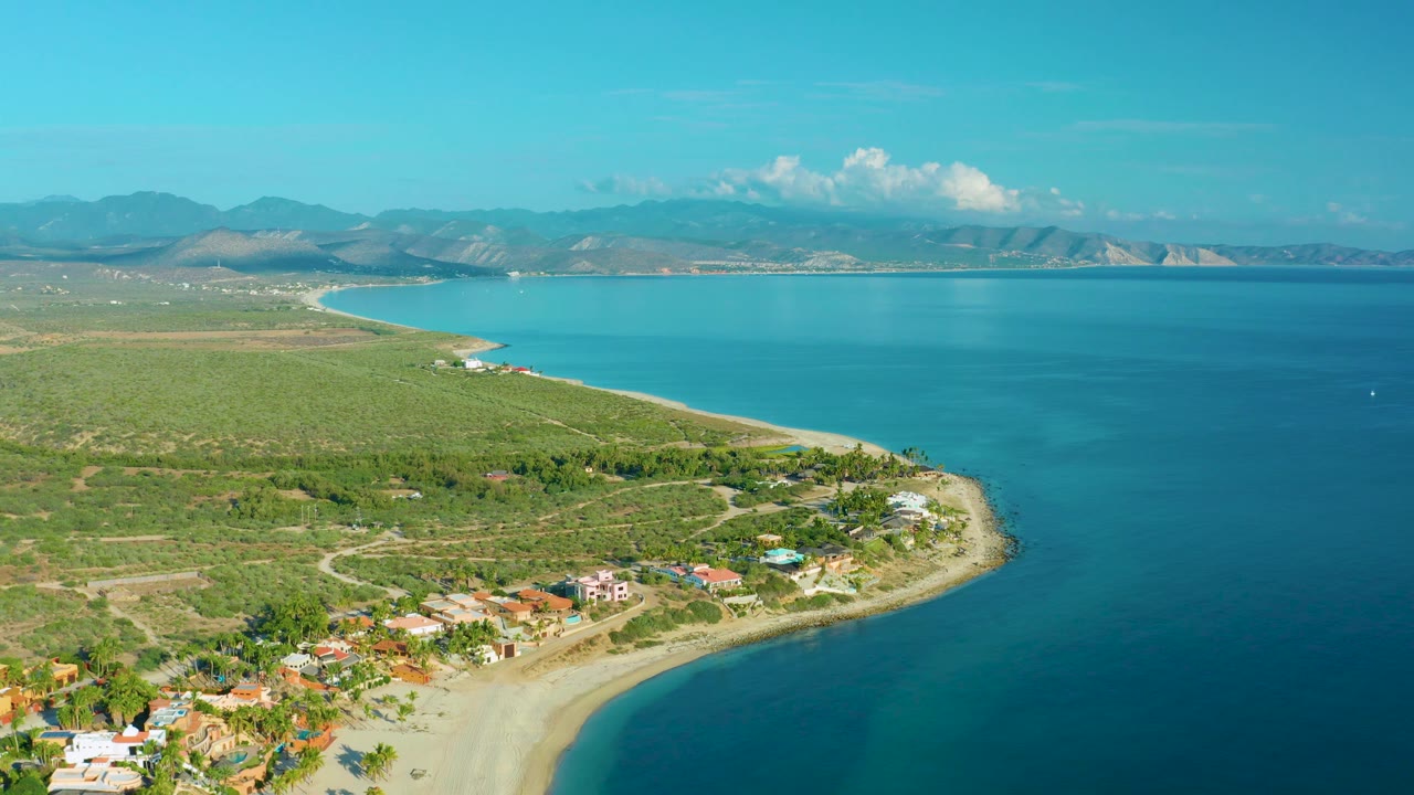 High coastal view of beach, scrubland, small buildings, bay water, and mountain backdrop.
