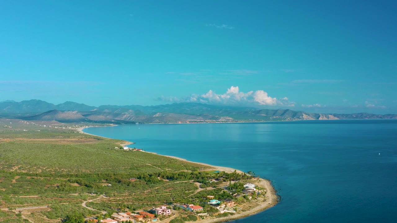 Opening aerial view over La Ribera, Baja California Sur, with green coastal fields, a curving shoreline, blue bay water, distant mountains, bright sky, and no visible people.
